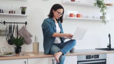 Asyalı bir kadın mutfak duvarında fotokopi alanı ile otururken dizüstü bilgisayar kullanıyor. Gündelik kıyafetlerle çekici bir kadın tele-çalışma sırasında esnek bir program izliyor..