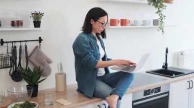Gündelik giyimli Japon kadın portresi. Parlak mutfakta iş yüzeyinde çıplak ayakla otururken bilgisayar tutuyor. Kendine güveni tam, gözlüklü, modern apartmanda ev tabanlı bir iş yapıyor..