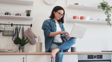 Gündelik giyimli Japon kadın portresi. Parlak mutfakta iş yüzeyinde çıplak ayakla otururken bilgisayar tutuyor. Kendine güveni tam, gözlüklü, modern apartmanda ev tabanlı bir iş yapıyor..