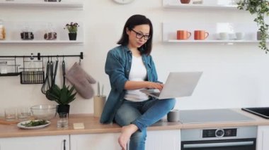 Asyalı bir kadın mutfak duvarında fotokopi alanı ile otururken dizüstü bilgisayar kullanıyor. Çekici bir kadın, günlük giysiler içinde, kameraya bakarken esnek bir program izliyor.