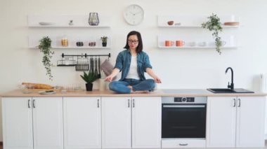 Barışçıl Asyalı kadın beyaz tasarımcı mutfak setinde otururken meditasyon yapıyor. Rahat bir dairede yaşam dengesini ve ahengi sağlamak için günlük giysiler giymiş ince bir kadın yoga yapıyor..