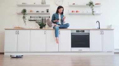 Robot toz toplayıcısı ıslak temizlik yaparken mutlu Asyalı kadın elinde modern akıllı telefonuyla mutfakta oturuyor. Koyu renk saçlı kadın internette sörf yapıyor ve ev işleri sırasında modern aletlerle dinleniyor..