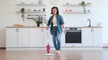Neşeli kadın, kablosuz kulaklıkla enerjik müzik dinlerken yerleri kablosuz makineyle süpürüyor. Aktif Asyalı kadın, evi iyi bir ruh haliyle temiz tutuyor. Geniş mutfağın arka planı.