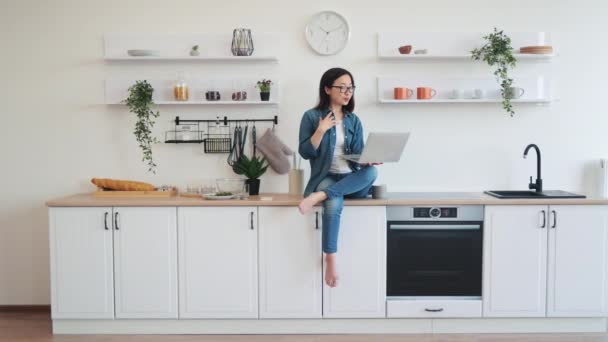 広々としたキッチンでノートパソコンのウェブカムを見ながら カジュアルな服で笑顔のブルネット女性 リモートワークの状況でアプリを介してビデオ通話を開始する光景で日本のプロフェッショナル — ストック動画