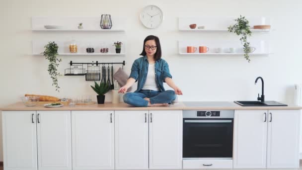 和平的亚洲女人坐在白色设计的厨房用具上沉思 身着休闲装的瘦小女性在舒适的公寓里练习瑜伽 以达到生活的平衡与和谐 — 图库视频影像