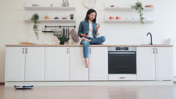 キッチンカウンターに足を横切って座っている間 ロボット掃除機を監視している若いアジアの女性 自宅でモダンなガジェットで簡単にクリーニング — ストック動画