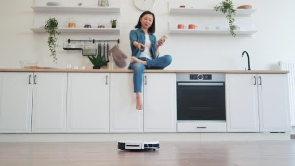 白いキッチンセットで休憩しながら 無線リモコンを介してロボット掃除機を接続する美しいアジアの女性 自宅でクリーニングプロセスを楽しんでいる手のスマートフォンを持つ若いブルネット — ストック動画