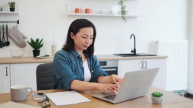 Mutfakta kalırken, rahat kıyafetli, taşınabilir dijital cihaz kullanan çekici bir kadının yan görüntüsü. Düşünceli Japon kadın araştırma projesi için online kütüphaneden kitap indiriyor..