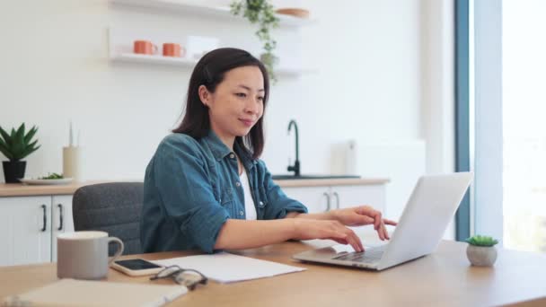 快適な部屋でデスクの表面にデジタルデバイスと文書をポーズする中国人女性の笑顔の肖像 デニム服の陽気な起業家は コンピュータを介してウェブページ上のデータを収集します — ストック動画