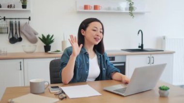 Çekici Asyalı kadın kişisel bilgisayarında online konferans sırasında el sallıyor. Koyu renk kısa saçlı genç bayan rahat dairesinde video görüşmesi yapıyor. Tasarımcı mutfağının arka planı.