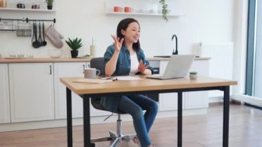 Aşırı neşeli Asyalı öğretim görevlisi, mutfaktaki ahşap masada otururken, akıllı teknolojiler vasıtasıyla çevrimiçi webinar sunuyor. Heyecanlı genç öğretmen yeni iş planıyla ilgili canlı yayın yapıyor..