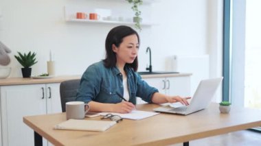 Modern mutfaktaki dizüstü bilgisayarlarda web sayfalarını kaydırırken kağıda odaklanmış Koreli yetişkin yazma bilgileri. Gündelik kıyafet giyen düşünceli kadın internet uygulaması aracılığıyla iş ağı hakkında bilgi topluyor.