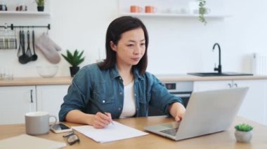 Modern laptop, kahve, bardak ve akıllı telefon ile ahşap masada otururken, Asyalı esmer, kağıt notlara bilgi yazıyor. Ev ofisi kavramı, online çalışma ve aletler.