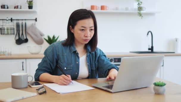 広々とした部屋でポータブルコンピュータの記事を読んでいる間 アジアの女性がノートを作るのを見る テストを書くためのオンライン教育リソースを使用してカジュアルウェアに焦点を当てた女性大人のテスト — ストック動画