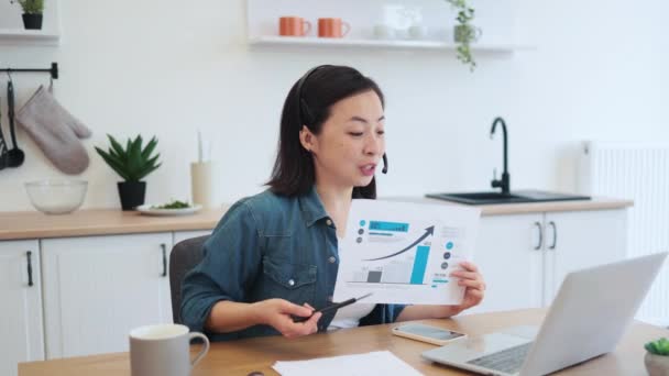 デニムウェアとヘッドセットのポジティブなアジアの女性は 屋内でのビデオ通話中に紙にグラフィック素材を示しています 自宅から遠隔で同僚と財務データプランを議論する楽しい従業員 — ストック動画