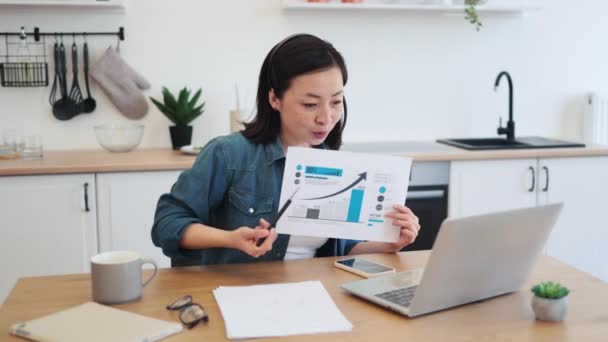 勤勉な女性は ヘッドセットの職場に座って 金融グラフにペンを指差しています 快適なキッチンインテリアでパーソナルノートパソコンでオンライン会議を持つ魅力的なアジアの女性 — ストック動画