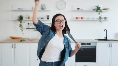 Evinde mikrofon yerine sahnede çırpıcıyla şarkı söylüyormuş gibi yapan rahat giysili güzel Asyalı kadın. Genç neşeli ev hanımı yemek pişirmeye başlamadan önce mutfak aletleriyle oynuyor..