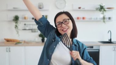 Evinde mikrofon yerine sahnede çırpıcıyla şarkı söylüyormuş gibi yapan rahat giysili güzel Asyalı kadın. Genç neşeli ev hanımı yemek pişirmeye başlamadan önce mutfak aletleriyle oynuyor..