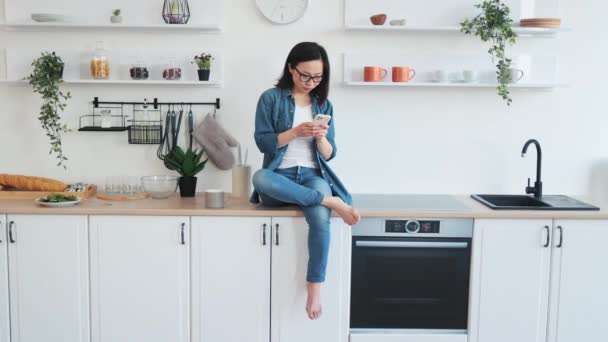 亚洲女人坐在白色宽敞厨房的台面上 使用智能手机的全景 无忧无虑的赤脚女士使用数码设备在烹饪网站上搜索新食谱 — 图库视频影像