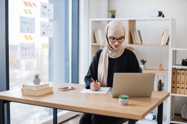 Günümüz iş yerinde dizüstü bilgisayar ekranına bakarken tesettürlü kadın kalemle not alıyor. Verimli Arap çalışanı kablosuz teknolojiler kullanarak şirketin gelişimi hakkında online araştırma yapıyor..