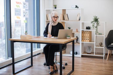 İş yerinde taşınabilir bilgisayarda yazı yazarken türban takmış güzel bir kadının tam boy görüntüsü. Ofis içinde e-ticaret yatırım stratejisi planlayan odaklı Müslüman girişimci.
