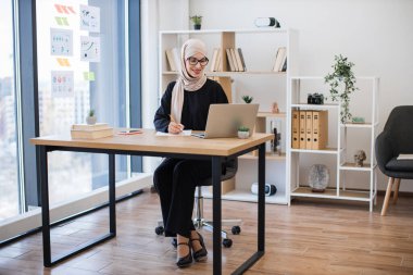 İş yerinde taşınabilir bilgisayarda yazı yazarken türban takmış güzel bir kadının tam boy görüntüsü. Ofis içinde e-ticaret yatırım stratejisi planlayan odaklı Müslüman girişimci.