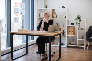 Çalışma saatleri boyunca ofis masasında hayal kurarken uzaktan bakan tesettürlü çekici Arap bayan. Dijital aygıt kullanılarak yıllık kurumsal etkinlik fikirlerini not alan dikkatli yönetici.