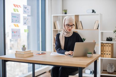 Başörtüsü takmış kendine güvenen genç bir çalışan modern ofiste masa şifresini içeren dizüstü bilgisayarın klavyesini açıyor. Yaratıcı iş yerinde arama motoru kullanarak markaları araştıran Müslüman kadın işçiler.