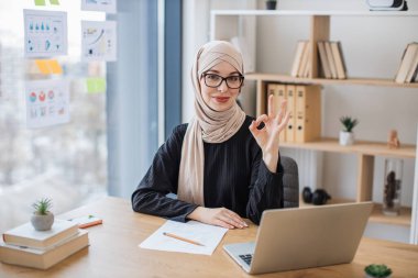 Çalışırken kablosuz cihaz arkasında poz veren gözlüklü çekici Arap kadın portresi. İşaret tamam.