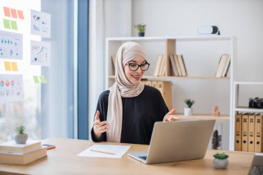 Geleneksel türbanlı gülümseyen iş kadını ofis masasında otururken bilgisayardan video mesajı alıyor. Planlanan online iş toplantısı sırasında neşeli Müslüman profesyonel meslektaşları selamlıyor.