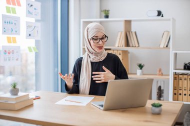 Geleneksel türbanlı gülümseyen iş kadını ofis masasında otururken bilgisayardan video mesajı alıyor. Planlanan online iş toplantısı sırasında neşeli Müslüman profesyonel meslektaşları selamlıyor.