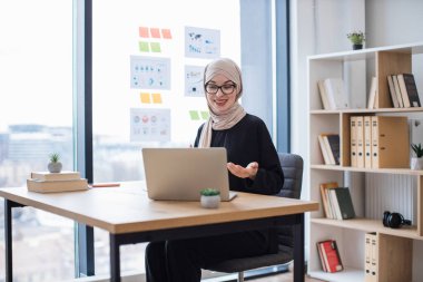 Başörtüsü takmış çekici bir kadın ofis masasında dizüstü bilgisayarlı web kamerasıyla sohbet ediyor. Projeyi yönetmek için meslektaşlar arasında görsel bağlantı kuran kendinden emin Müslüman yönetici.