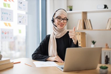 Ofis masasında bilgisayarla otururken kulaklık takan mutlu yetişkin bir kadın. Gülümseyen Müslüman göz giyimi danışmanı çevrimiçi sohbetten sonra başarılı bir anlaşmayı onaylıyor.