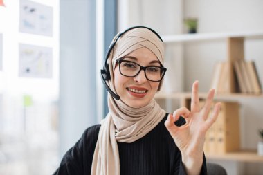 Ofis masasında bilgisayarla otururken işaret gösteren kulaklıklı mutlu yetişkin bir kadın. Gülümseyen Müslüman göz giyimi danışmanı çevrimiçi sohbetten sonra başarılı bir anlaşmayı onaylıyor.