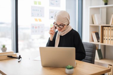 Çekici bir Arap iş yerindeki dizüstü bilgisayarda web sayfalarını kaydırırken cep telefonuyla önemli bir arama yapıyor. Ofisteki dijital aygıtlar aracılığıyla başlangıç alışverişi tesettüründeki mutlu girişimci.