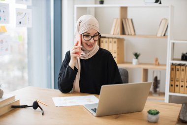 Geleneksel Müslüman başörtülü neşeli bayan dijital ekrandan haberleri okurken akıllı telefondan konuşuyor. Deneyimli çalışanlar iş yerlerindeki şirket verimliliğini artırma fikrini ortaya attılar..
