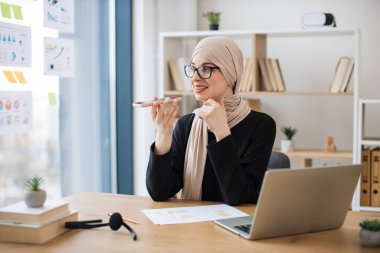 Geniş iş iş yerinde telefon görüşmesi sırasında hoparlör uygulaması kullanarak geleneksel türbanlı gülümseyen bayan. Gözlüklü neşeli Müslüman profesyonel ofiste çalışma saatlerinde danışmanlık yapıyor..