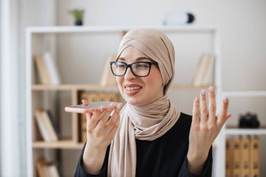 Geniş iş iş yerinde telefon görüşmesi sırasında hoparlör uygulaması kullanarak geleneksel türbanlı gülümseyen bayan. Gözlüklü neşeli Müslüman profesyonel ofiste çalışma saatlerinde danışmanlık yapıyor..