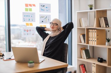 Geleneksel türbanlı çekici Müslüman kadın ofis sandalyesinde kolları başının arkasında dinleniyor. Ofisteki iş ortaklarıyla müzakere sürecinden sonra kaygısız profesyonel stres giderme.