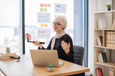 Akıllı telefonlu, selfie çubuklu güzel Müslüman kadın ofis içi video bloğu için video içeriği kaydediyor. Taraftarlar için iş hayatı dengesi konusunu kapsayan geleneksel türbanlı keyifli kadın.