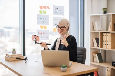 Güzel müslüman kadın, akıllı telefonlu, selfie çubuklu, video içeriğini kaydeden ofis içi video bloğu, baş parmağını gösteriyor.
