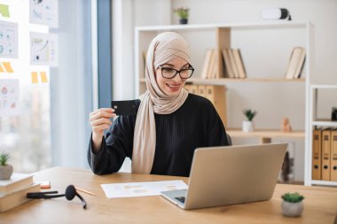 Ofisin içinde bilgisayar ekranının arkasında otururken siyah kredi kartı tutan başörtüsü takan gülümseyen genç bir kadın. Rahat Müslüman çalışan internet uygulaması kullanarak online bankacılık işlemi yapıyor.