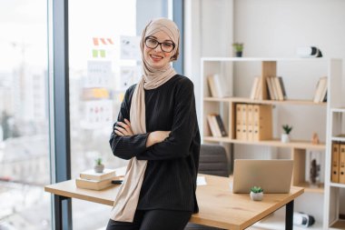 Elleri göğsünde katlanmış, ahşap masanın kenarında oturan, göz giysisi giymiş gülümseyen Arap yetişkin portresi. Bej tesettürlü neşeli iş kadını modern işyerinden başarılı bir şirket yönetiyor..