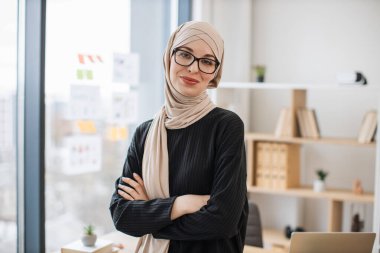 Elleri göğsünde katlanmış, ahşap masanın kenarında oturan, göz giysisi giymiş gülümseyen Arap yetişkin portresi. Bej tesettürlü neşeli iş kadını modern işyerinden başarılı bir şirket yönetiyor..