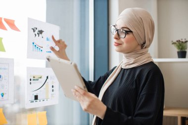 Dijital tabletli çekici Arap bayan iş yerindeki şema ve çizelgeleri kullanarak finansal istatistikleri inceliyor. Akıllı aygıttaki bilgileri kağıt raporuyla eşleştiren yetkili veri analisti.