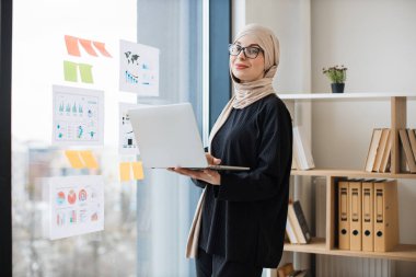 Başörtülü güzel bir kadın ofis penceresinin yanında portatif bilgisayarla dikiliyor. Düşünceli Müslüman profesyonel personel toplantısı sırasında online sunuma hazırlanıyor..