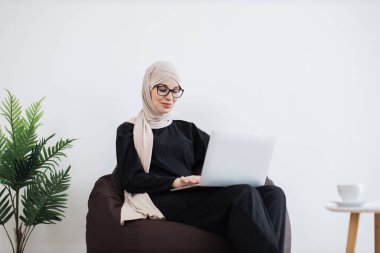 Portatif bilgisayar kullanırken yeni fikirler üzerine düşünen tesettürlü çekici Arap kadınına yakından bakın. Fotokopi alanının yakınındaki puf minderinde dinlenirken stratejik hedefler geliştiren etkili bir uzman..