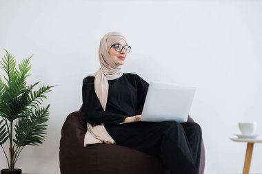 Portatif bilgisayar kullanırken yeni fikirler üzerine düşünen tesettürlü çekici Arap kadınına yakından bakın. Fotokopi alanının yakınındaki puf minderinde dinlenirken stratejik hedefler geliştiren etkili bir uzman..