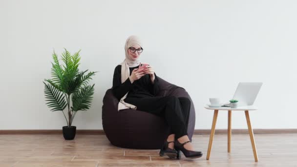 ハッピーイスラム教徒の女性は コーヒーテーブルの近くのポーフ椅子で休んでいる間 携帯電話を保持しています 笑顔のフリーランサーは コピースペースで内部のクライアントや同僚からのメッセージに絶えず答えます — ストック動画