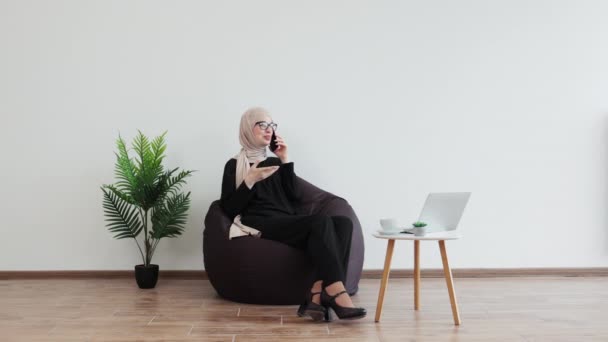 ビーンバッグでリラックスしながら 携帯電話で話すヘッドスカーフと正式な服の陽気なイスラム教徒の女性 コピースペースで職場の快適さで効果的な起業家リーディングビジネス会話 — ストック動画